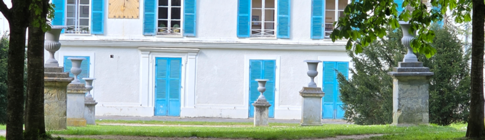 allée bordée de 6 vasque sur des socle, face à un bâtiment blanc avec des volets bleu turquoise