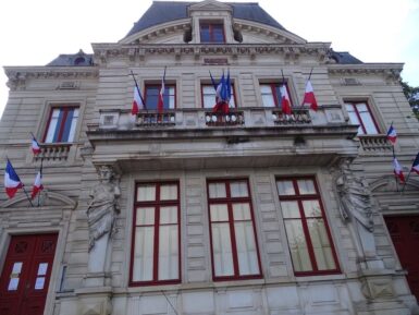 Façade XIXème de deux étages avec toit mansardé et lucarnes. Deux cariatides en pierre supportent un balcon. Des drapeaux français flottent sur la façade.